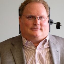 Head shot of Tim Fox, white man in sports coat; wheelchair headrest is visible.