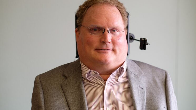 Head shot of Tim Fox, white man in sports coat; wheelchair headrest is visible.