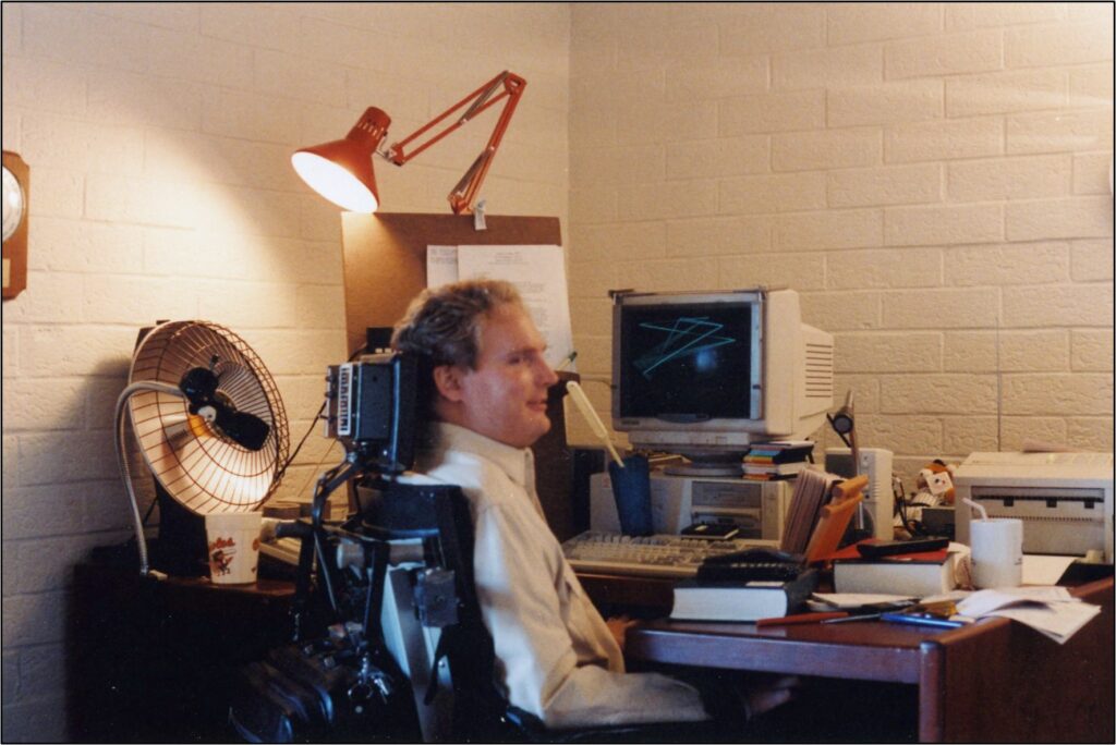 Tim (white man, blond hair, shirt & tie, in wheelchair by crowded desk with old-style monitor and desk lamp.